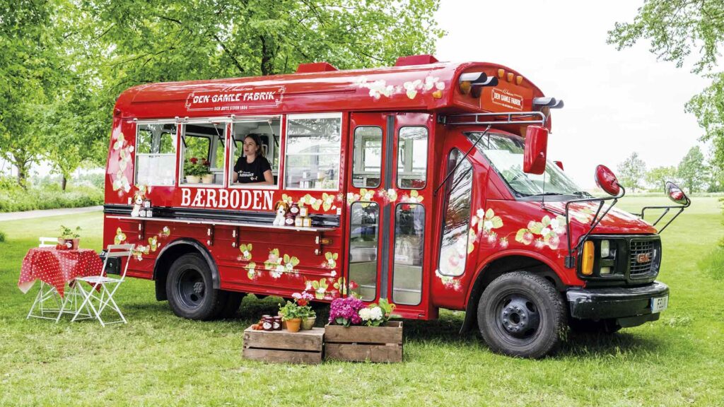 Et udendørsbillede af Den Gamle Fabriks bærbod, hvor der står to kasser med blomster og et havebord foran, mens en pige kigger ud af et åbent vindue i bærboden.
